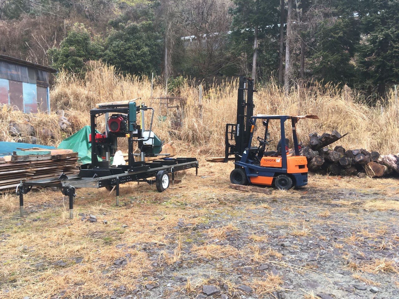 土場に置かれた移動式製材機（緑）とフォークリフト（トヨタ）。左に板を積んだ山、右に丸太