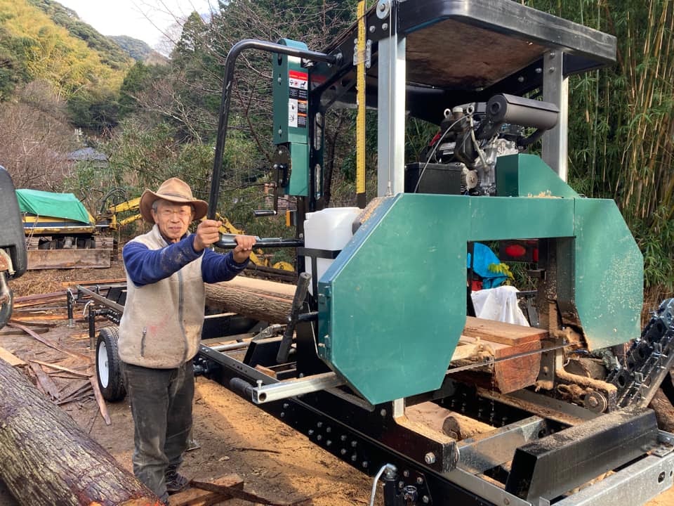 移動式製材機のそばに立つ人。機械に丸太が載っている